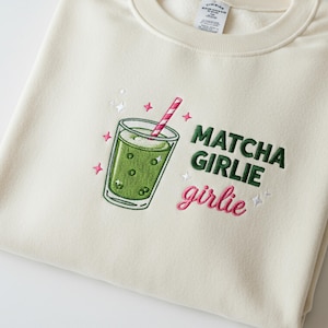 May include: Cream-colored sweatshirt with an embroidered matcha drink design. The design includes a green drink in a glass with a pink and white striped straw, and the words "Matcha Girlie girlie" in green and pink.