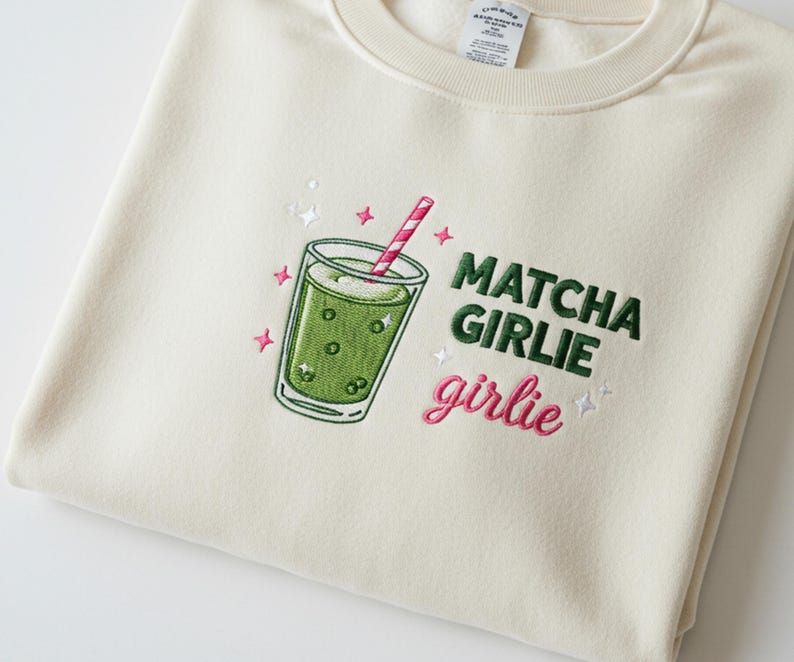May include: Cream-colored sweatshirt with an embroidered matcha drink design. The design includes a green drink in a glass with a pink and white striped straw, and the words "Matcha Girlie girlie" in green and pink.