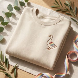 Pode incluir: Uma camisola bege dobrada com gola redonda e um pequeno desenho de ganso bordado na frente. O ganso tem uma asa colorida com as cores do arco-íris. Uma fita de arco-íris está enrolada perto e vegetação no fundo.