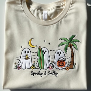 May include: Off-white t-shirt with a cartoon beach scene featuring three ghost characters. One ghost holds pizza, another a surfboard, and the third a jack-o'-lantern. A crescent moon, stars, and palm tree are also shown. The text "Spooky & Salty" is below.
