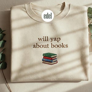 May include: A cream-coloured sweatshirt with the text "will yap about books" embroidered in brown. A stack of books is embroidered below the text.