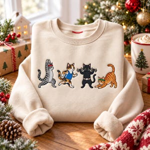 May include: A beige crewneck sweatshirt featuring a graphic print of four cats exercising. One gray tabby cat does yoga, a calico cat runs, a black cat lifts dumbbells, and an orange tabby cat stretches. Each cat wears a headband or athletic gear.