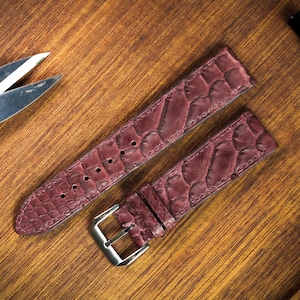 Könnte beinhalten: Burgunderfarbene Uhrenarmbänder mit einem strukturierten, schuppenartigen Muster. Die Armbänder bestehen aus einem geschmeidigen Material und verfügen über eine silberfarbene Schnalle. Die Uhrenarmbänder werden auf einer Holzoberfläche präsentiert.