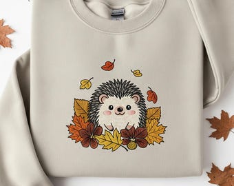 May include: Beige sweatshirt featuring an embroidered hedgehog surrounded by autumn leaves and flowers in shades of orange, yellow, and brown. The hedgehog has black quills and a smiling face. A cozy fall-themed design.