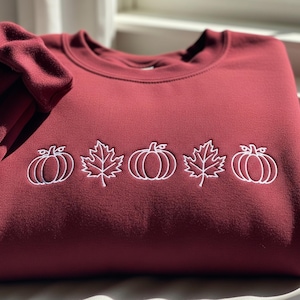 Peut inclure: Un sweat-shirt bordeaux orné de broderies blanches de citrouilles et de feuilles. Le motif comprend deux citrouilles, une feuille, une autre citrouille, une feuille et une dernière citrouille. Le sweat-shirt est en matière douce, parfait pour l'automne.