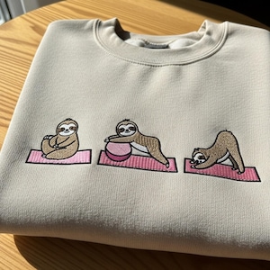 May include: Beige sweatshirt featuring an embroidered design of three sloths in yoga poses on pink mats. The sloths are brown and cream-colored. The sweatshirt is made of a soft, comfortable material.