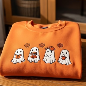 Puede incluir: Sudadera naranja con un diseño bordado de cuatro fantasmas de dibujos animados. Cada fantasma está representado con un libro o una taza, con hojas de arce encima. La sudadera está doblada sobre una superficie de madera.