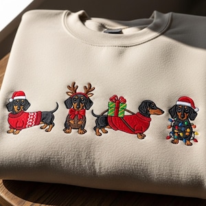 May include: A beige sweatshirt featuring an embroidered design of four dachshunds in festive holiday attire. Each dog is dressed in a Christmas sweater, Santa hat, reindeer antlers, or wrapped in Christmas lights. The design is centered on the front of the folded sweatshirt.