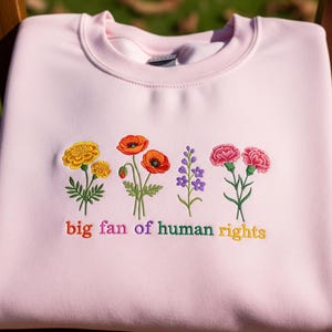Op de afbeelding: Een lichtroze sweatshirt met geborduurde bloemen in geel, oranje, paars en roze. De tekst "big fan of human rights" is onder de bloemen geborduurd in regenboogkleuren. De sweatshirt is op een houten oppervlak gevouwen.