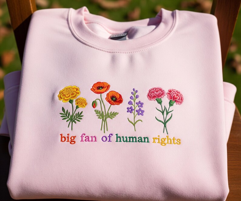 Op de afbeelding: Een lichtroze sweatshirt met geborduurde bloemen in geel, oranje, paars en roze. De tekst "big fan of human rights" is onder de bloemen geborduurd in regenboogkleuren. De sweatshirt is op een houten oppervlak gevouwen.