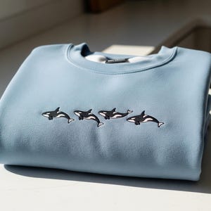 May include: A pale blue crew neck sweatshirt, folded and stacked. It has an embroidered design of four black and white orcas across the chest. The sweatshirt is made of a soft material. The garment is folded neatly.