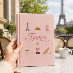 May include: A light pink hardcover book with embroidered icons including the Eiffel Tower, a croissant, a perfume bottle, a coffee cup, macarons, a baguette, a piece of cheese, and a fleur-de-lis, with the word France written in cursive script.