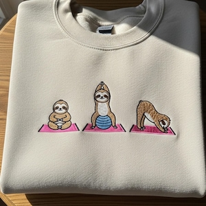 May include: A beige sweatshirt featuring embroidered sloths in yoga poses on pink mats. The sloths are in various positions, including seated, upward salute, and downward-facing dog. The sweatshirt is folded neatly on a wooden surface.
