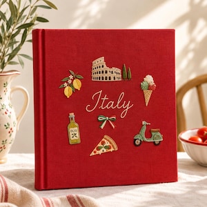 May include: A square red fabric-covered book with the word Italy embroidered in white script in the center. Surrounding the text are embroidered icons including a lemon branch, the Colosseum, an ice cream cone, a bottle of oil, a bow, a pizza slice, and a green scooter.