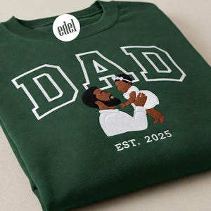 Puede incluir: Una sudadera verde oscuro con texto bordado en blanco que dice "DAD" y "EST. 2025". La sudadera presenta una imagen bordada de un padre y una hija negros.