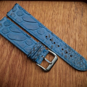 Op de afbeelding: Blauwe horlogeband gemaakt van getextureerd leer, met een schubbenpatroon en blauwe stiksels. De band heeft een zilverkleurige gesp en wordt getoond op een houten oppervlak.