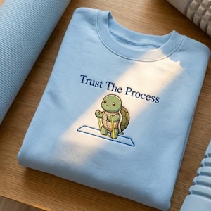 Op de afbeelding: Lichtblauw sweatshirt met de geborduurde tekst "Trust The Process" en een cartoon schildpad met trainingsbanden. De schildpad staat op een blauwe mat. Het sweatshirt is opgevouwen.