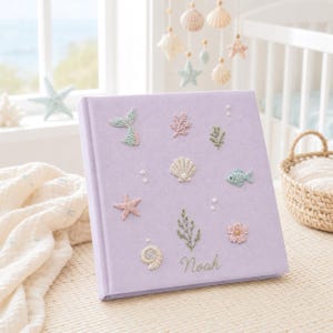 May include: A square, light purple velvet photo album featuring embroidered ocean-themed motifs including a mermaid tail, starfish, seashell, fish, and coral. The name Noah is embroidered in cursive script at the bottom right corner.