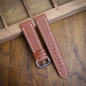 May include: Brown leather watch strap with white stitching and a silver buckle. The strap is shown on a wooden surface, highlighting the texture and color of the leather. The watch band is a classic design.