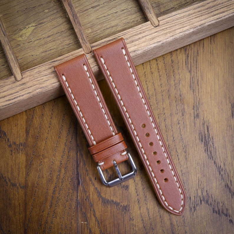 May include: Brown leather watch strap with white stitching and a silver buckle. The strap is shown on a wooden surface, highlighting the texture and color of the leather. The watch band is a classic design.