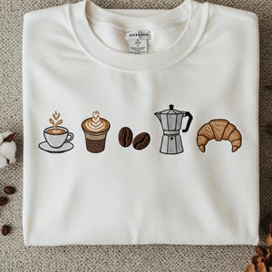 Peut inclure: Sweat-shirt blanc avec broderies sur le thème du café. Il présente une tasse de café, un latte, des grains de café, une cafetière italienne et un croissant. Les motifs sont dans les tons de marron, beige et noir. Le sweat-shirt a un col rond.