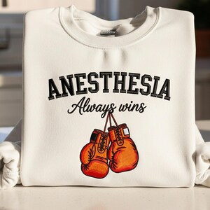 Könnte beinhalten: Cremefarbenes Sweatshirt mit dem Text "ANESTHESIA Always wins" in schwarzer Schrift. Darunter zwei orangefarbene Boxhandschuhe. Das Sweatshirt liegt auf einer weißen Oberfläche.
