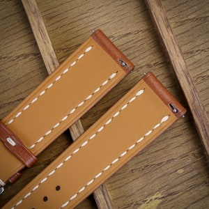 May include: Close-up of two tan leather watch straps with white stitching and brown edges. The straps are on a wooden surface. The straps have metal spring bars for attachment to a watch case.