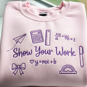 以下が含まれることがあります： 紫色の刺繍グラフィックとテキストが施された、薄いピンク色のスウェットシャツ。 デザインには、紙飛行機、電卓、定規、「Show Your Work」というフレーズが含まれています。 追加の数学記号とリボンも表示されています。