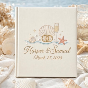 May include: A square off-white fabric-covered wedding album featuring gold embroidered designs of a seashell, two interlocking rings, a champagne flute, and a starfish. The text reads Harper & Samuel and March 27, 2028 in gold script.