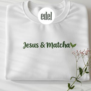 May include: White sweatshirt featuring the text "Jesus & Matcha" in green script, with a green leaf graphic. The sweatshirt has a crew neck and long sleeves. A sprig of pink flowers is in the lower right corner. The brand logo is in a white circle.