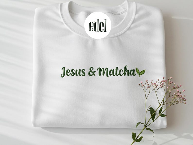 May include: White sweatshirt featuring the text "Jesus & Matcha" in green script, with a green leaf graphic. The sweatshirt has a crew neck and long sleeves. A sprig of pink flowers is in the lower right corner. The brand logo is in a white circle.