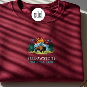 Op de afbeelding: Een bordeauxrood sweatshirt met een geborduurd ontwerp met een bergketen, een zon, een bizon en de tekst "EST. 1872 YELLOWSTONE NATIONAL PARK".
