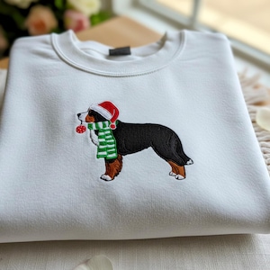 Pode incluir: Camisola branca com um bordado de um boiadeiro bernês usando um gorro de Papai Noel, um cachecol listrado verde e branco e segurando um enfeite vermelho. O cão é preto, marrom e branco. A camisola está dobrada.