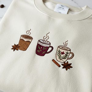 May include: A cream-coloured sweatshirt with embroidered images of three steaming mugs of coffee or tea. The mugs are brown, burgundy, and cream-coloured, with star anise and cinnamon sticks. The design evokes a cosy, warm beverage theme.