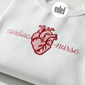 Może przedstawiać: Biała bluza z haftem w kolorze różowym i czerwonym przedstawiającym serce i napis "cardiac nurse".
