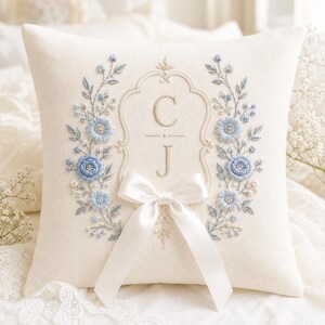 May include: A square off-white throw pillow featuring intricate embroidery of blue floral wreaths flanking a central ornate frame. The letters C and J are stitched in the center. A smooth white satin ribbon bow is attached at the bottom.