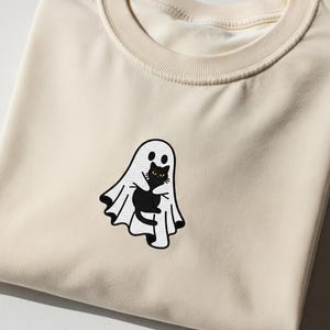 May include: A folded cream-coloured sweatshirt with an embroidered design. The design features a white ghost hugging a black cat with yellow eyes. A Halloween-themed garment. The sweatshirt is folded, showing the design on the front.