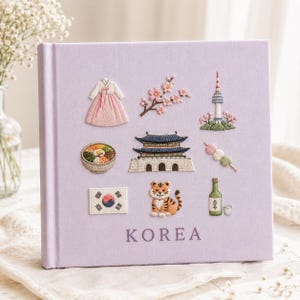 Op de afbeelding: Een vierkant, lichtpaars met stof bekleed boek met geborduurde iconen, waaronder een hanbok, kersenbloesems, de N Seoul Tower, een kom bibimbap, een paleis, dango, de Zuid-Koreaanse vlag, een tijger en een fles soju met het woord KOREA.