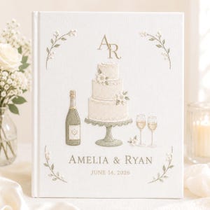 Peut inclure: Un livre d'or recouvert de tissu blanc avec des broderies dorées et crème. Le motif représente un gâteau de mariage à trois étages, une bouteille de champagne, deux verres et des brins de fleurs. Texte : AR, AMELIA & RYAN, JUNE 14, 2026.
