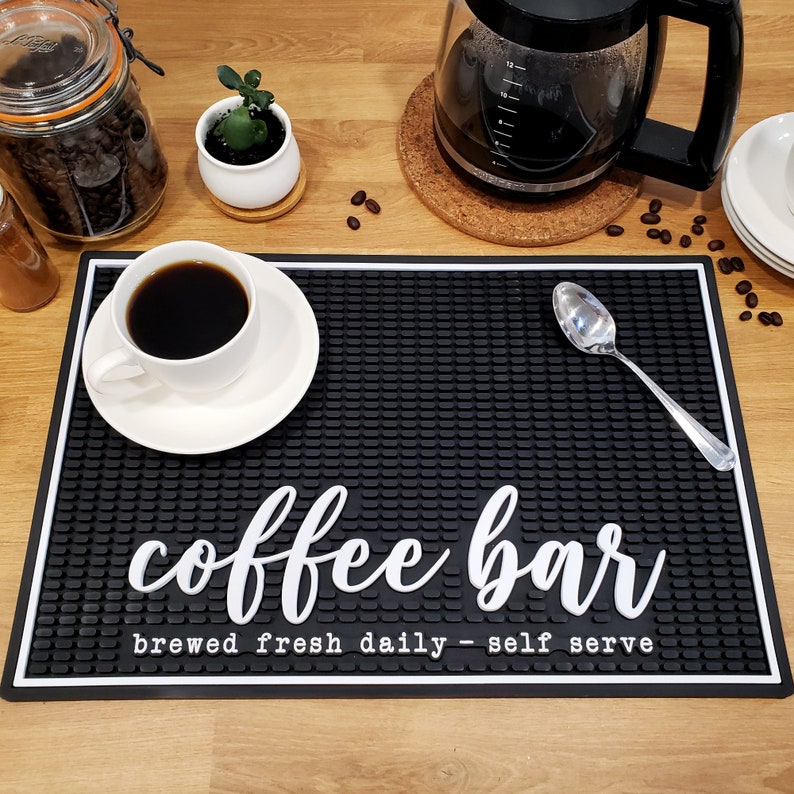 May include: Black rubber coffee bar mat with white text that reads "coffee bar brewed fresh daily - self serve". A white cup of coffee and a white saucer are on the mat. A silver spoon is on the mat.