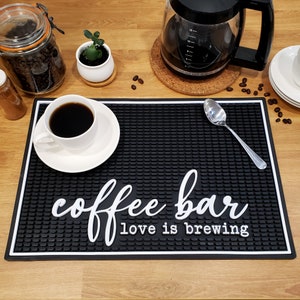 May include: Black and white coffee bar mat with the text "coffee bar love is brewing". A white coffee cup with black coffee is on a white saucer on the mat. A silver spoon is on the mat.
