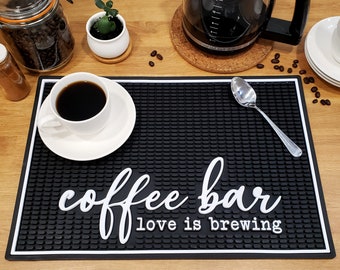 May include: Black and white coffee bar mat with the text "coffee bar love is brewing". A white coffee cup with black coffee is on a white saucer on the mat. A silver spoon is on the mat.