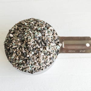 May include: A stainless steel measuring cup filled with a mixture of small, coarse, multi-colored gravel stones in shades of black, light gray, and pale green. The handle of the cup is clearly marked with the text 480ml and 2CUP.