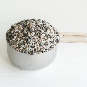 May include: A stainless steel measuring cup filled with a mix of small, irregularly shaped pebbles. The pebbles are a mix of gray, black, and light green colors. The cup is on a white wooden surface.