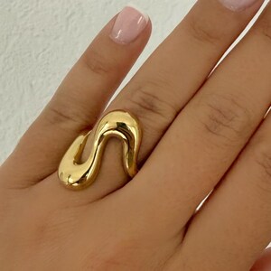May include: A polished gold ring featuring a continuous, fluid, and wavy design that wraps around the finger. The metal surface is smooth and reflective, creating a series of soft, organic curves that form an abstract, interconnected shape.