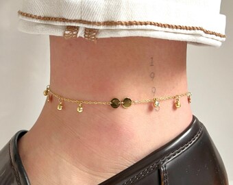 May include: A delicate gold anklet with small dangling charms and disc-shaped accents. The anklet is worn on an ankle, paired with a black leather shoe and white denim jeans.
