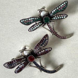 Może przedstawiać: Dwie broszki w kształcie ważki z korpusami z metalu w kolorze srebrnym. Jedna ma zielone i różowe akcenty, druga czerwone i niebieskie. Każda ma główkę przypominającą perłę i centralny kamień szlachetny.