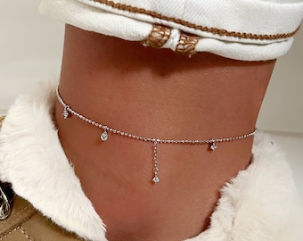 May include: A delicate silver anklet with dangling charms and small, sparkling accents. The anklet is worn on an ankle, paired with a tan sneaker with a white faux fur lining and white jeans.
