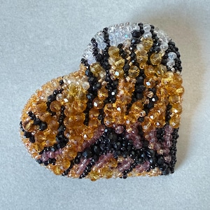May include: A heart-shaped beaded brooch with a gradient of colors. The brooch features a mix of black, amber, and clear beads, with a touch of pink. The beads are arranged to create a textured surface.