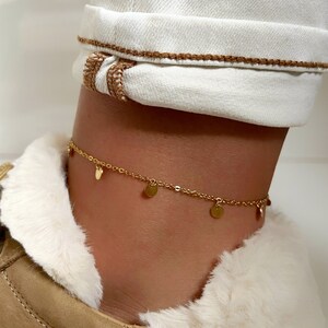 May include: A delicate gold-colored anklet with small, round charms. The anklet is worn on an ankle, paired with white pants and a tan boot with a faux fur lining. The charms are evenly spaced along the chain.